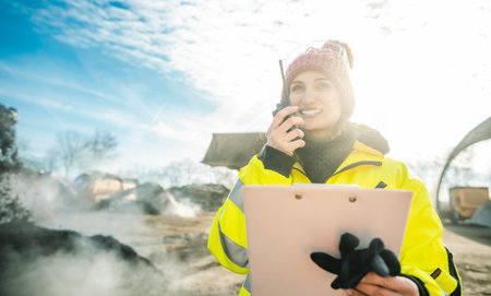 Manager in biomass and landfill operation using her radio in front of machines, clipboard in hand