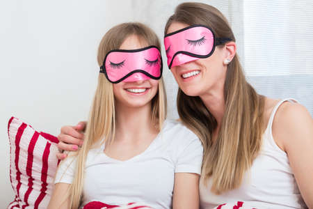 Mother and daughter wearing pink sleeping masks in bedの写真素材