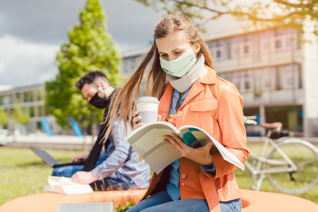 Woman student on college campus learning wearing face mask while working on her laptopの写真素材