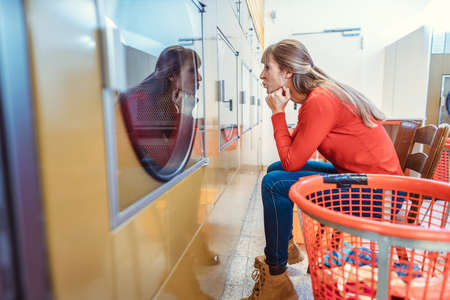 A woman waits sitting in front of a washing machine in a laundromatの写真素材