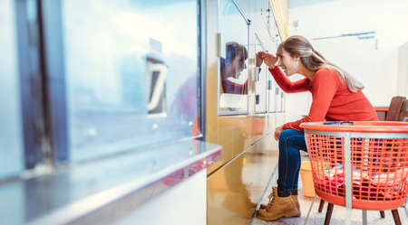 A woman waits sitting in front of a washing machine in a laundromatの写真素材