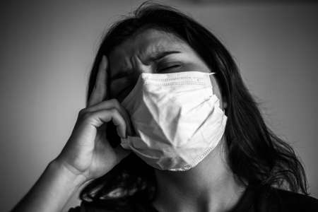 Portrait of a young woman wearing medical sterile mask, having a strong painful headache. Dramatic black and white closeup of a girl being protected from coronavirus. Quarantine stay home conceptの写真素材