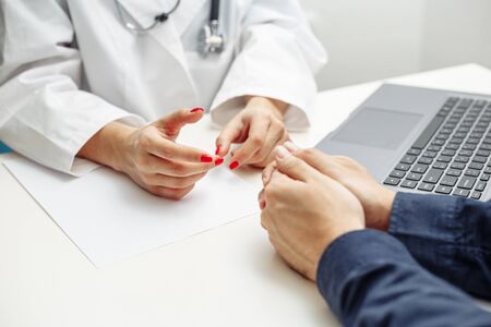 Closeup of a female doctor hands with a green pen making notes on a laptop and filling in diagnosis on a paper. Discussing treatment with a patient. Coronavirus test and prevention conceptの写真素材