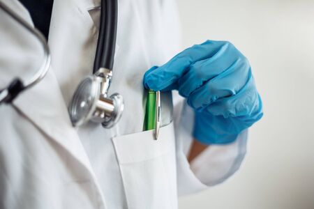 Closeup of a woman doctor wearing white medical gown and blue gloves, stethoscope. Female nurse taking a green pen from her pocket. Coronavirus, covid-19 and sickness prevention conceptの写真素材