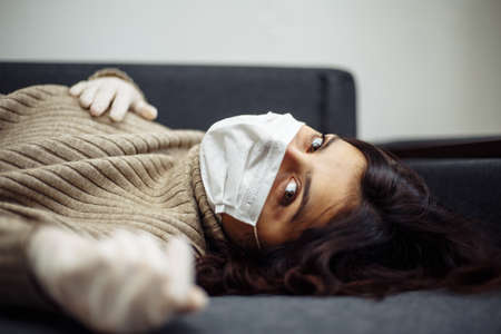 Young woman bored sitiing home during quarantine due to coronavirus pandemia. Beautiful girl stays home wearing medical mask and gloves laying on a sofa. Covid-19 epidemia worldwide conceptの写真素材