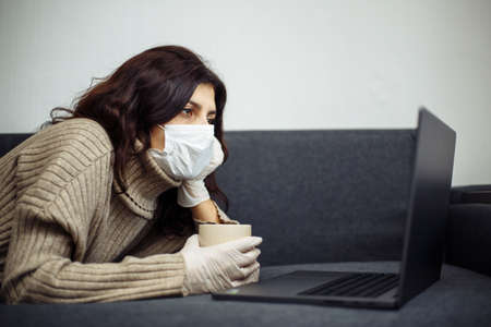 Young woman working from home during quarantine due to coronavirus pandemia. Beautiful girl stays home wearing medical mask and gloves and typing on a laptop. Covid-19 epidemia worldwide conceptの写真素材