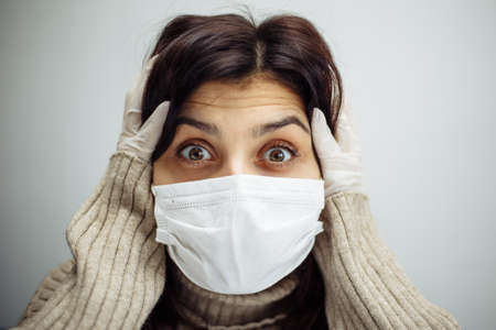 Portrait of a young woman holds hands on her head and has to stay home during quarantine due to coronavirus pandemia. Beautiful girl wearing medical mask and gloves. Covid-19 epidemia conceptの写真素材