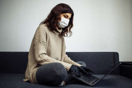Young woman working from home during quarantine due to coronavirus pandemia. Beautiful girl stays home wearing medical mask and gloves and typing on a laptop. Covid-19 epidemia worldwide conceptの写真素材