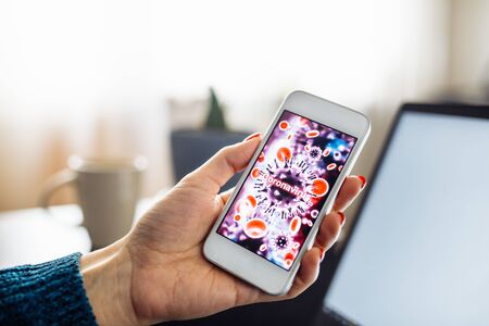 Closeup of a mobile cell phone of a young woman staying home and leadling business from distance on a freelance basis. Laptop, cup and flower in the room on the background. Coronavirus sign on the screenの写真素材