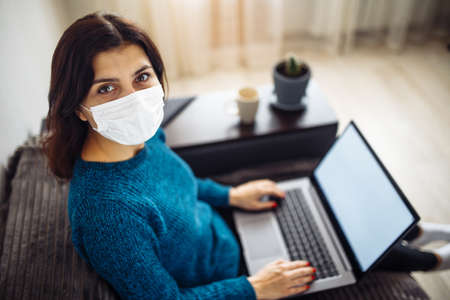 Businesswoman stays home and works during coronavirus epidemia quarantine. Female worker wearing a medical mask and typing on a laptop. Covid-19 pandemia spread prevention conceptの写真素材