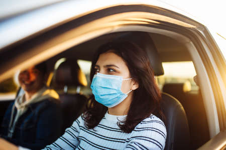 A girl drives a car with a passanger wearing medical mask during coronavirus pandemic quarantine. Woman taxi driver stuck in a traffic jam. Stay safe and healthcare conceptの写真素材