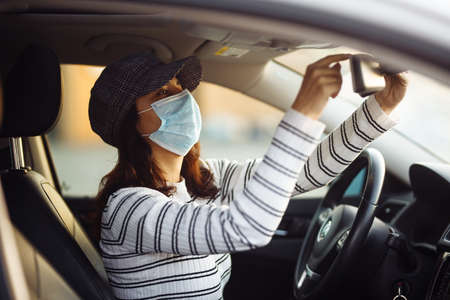 Woman car taxi driver adjusts backview mirror wearing medical sterile mask during pandemic...