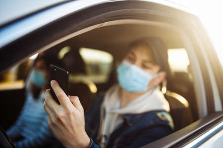 A man wearing a medical mask holds a mobile cell phone in his hand while driving a car. Boy driver checking coronavirus world news in a traffic jam. Isolation and healthcare during quarantine conceptの写真素材
