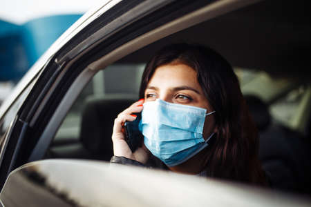 Woman wearing a medical sterile mask in a taxi car on a backseat looking out of window talking on the phone. Girl passenger waiting in a traffic jam during coronavirus quarantine. Healthcare conceptの写真素材
