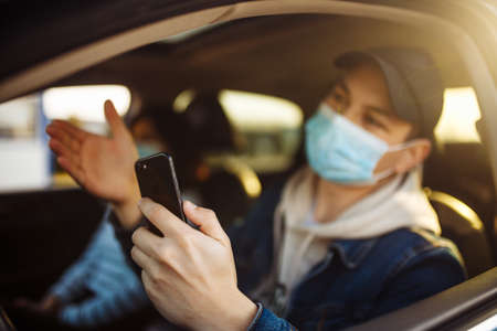 A man wearing a medical mask holds a mobile cell phone in his hand while driving a car. Boy driver...