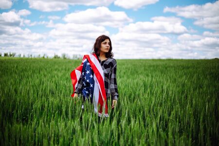 Young woman wrapped in an american flag on the green wheat field. Patriotic holiday celebration....