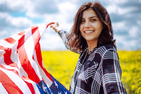 Patriotic young woman wrapped in an american flag on a beautiful field with yellow flowers. USA...