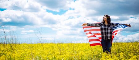 Patriotic young woman wrapped in an american flag on a beautiful field with yellow flowers. USA independence day 4th of July celebration. Summer holidays conceptの写真素材