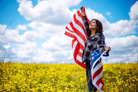 Patriotic young woman wrapped in an american flag on a beautiful field with yellow flowers. USA independence day 4th of July celebration. Summer holidays conceptの写真素材