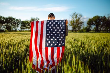 Young man wearing green shirt and cap stands wrapped in the american flag at the green wheat field....