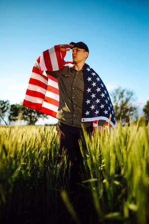 Young man wearing green shirt and cap stands wrapped in the american flag at the green wheat field. Patriotic boy celebrates usa independence day on the 4th of July with a national flag in his handsの写真素材