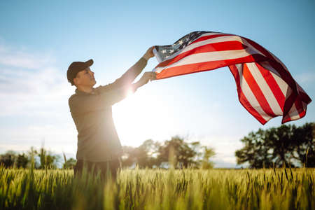 Young man wearing green shirt and cap lets the american flag fly on the wind at the green wheat field. Patriotic boy celebrates usa independence day on 4th of July with a national flag in his handsの写真素材
