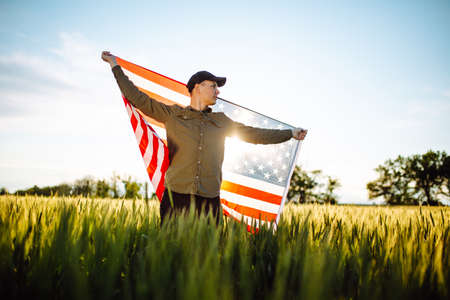Young man wearing green shirt and cap lets the american flag fly on the wind at the green wheat...