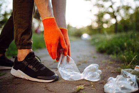 Young man volunteer picks up trash at the park. Plastic bottle is being collected by the man wearing orange gloves. Pollution, ecology, environment concept.の写真素材