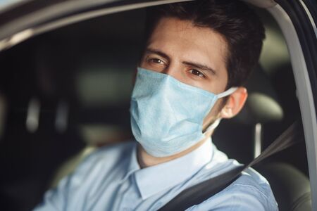 Closeup portrait of young man sits in the car wearing sterile medical mask. Boy taxi driver works...