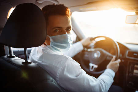 Young man taxi driver steers the car during coronavirus pandemic wearing sterile medical mask. A boy drives a car through the traffic on the sunset. Social distance and health care conceptの写真素材