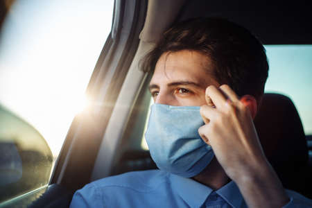 Young businessman takes a taxi and looks out of the car window adjusting sterile medical mask. Man sits on the back seat of taxi and takes a ride during coronavirus pandemic. Social distance conceptの写真素材