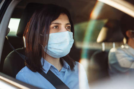 Young girl passenger takes a ride by taxi during the coronavirus pandemic quarantine. Woman looks out of the car window wearing sterile medical mask. Social distance and health safety conceptの写真素材