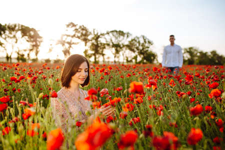 Beautiful young woman enjoys sitting among red poppy flowers on the field on the sunset. A girl feels harmony with nature and touches the plants. Love, peace and satisfaction conceptの写真素材