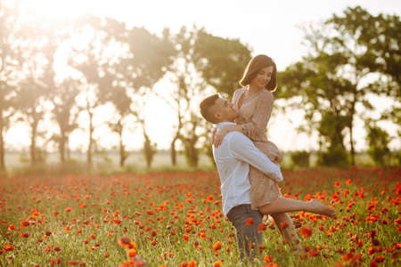 Young romantic loving married couple is standing among the red poppy flowers on the field on the beautiful sunset. Attractive boy and tender girl hug and kiss feeling happy. Love family conceptの写真素材