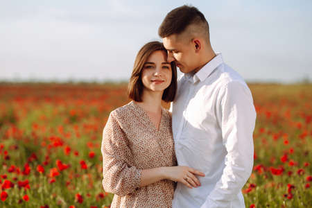 Young romantic loving married couple is standing among the red poppy flowers on the field on the beautiful sunset. Attractive boy and tender girl hug and kiss feeling happy. Love family conceptの写真素材