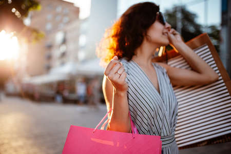 Happy girl stands near boutique shop holding a few bags with purchases. Young woman is glad to buy new clothes during sales season for a reasonable price. Shopping, discount, spend money conceptの写真素材