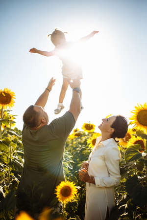 Happy family having fun in the field of sunflowers. Father and mother throw their daughter in the air and smile. Girl likes playing with her parents. Summer season, freedom, family value conceptの写真素材