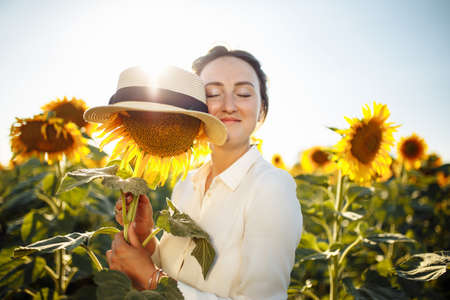 Young woman hugs a single sunflower in a hat on the background of a field of sunflowers on a sunny summer day. Joy, easy, happiness, freedom concept.の写真素材
