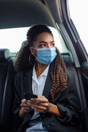 Young business woman in a mask checking her mobile cell phone on a backseat of a taxi during...