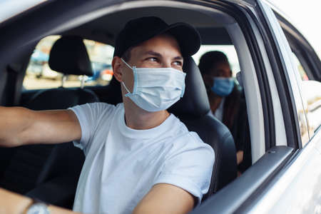 Young taxi driver looks out of a car's window while driving through the city with a passanger wearing a medical mask. Business trips during pandemic, new normal and coronavirus travel safety conceptの写真素材