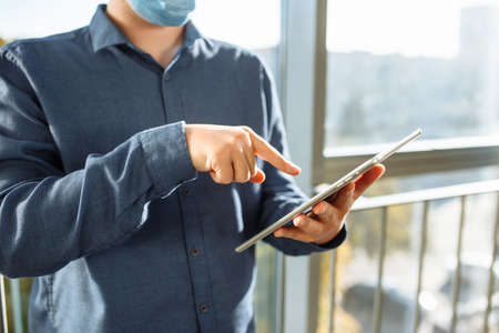 Young man with tablet in his hands standing at the office corridor wearing protective medical mask....
