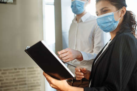 Two collegues discuss business matters at the corridor of the office wearing medical masks to...