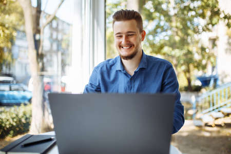 Cheerful businessman in a blue shirt working from a cafe on his laptop. Young man leads distance business with a cup of coffee. Remote job conceptの写真素材