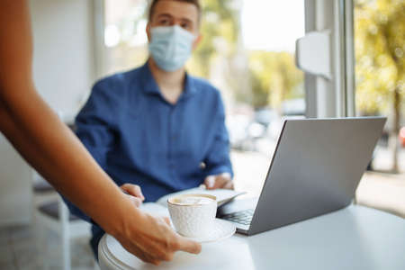 Waiter brings a cup of coffee to a young businessman wearing a sterile medical mask working from a...