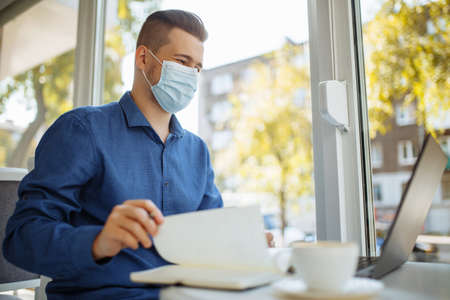Young businessman wearing a medical mask sits in a cafe with a laptop working and taking notes....