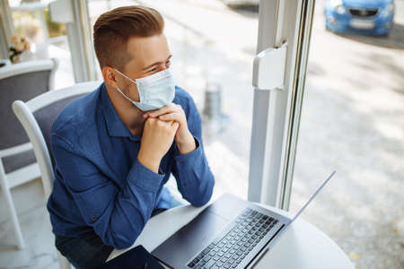 Young businessman wearing a medical sterile mask sits with a laptop in a cafe working remotely....