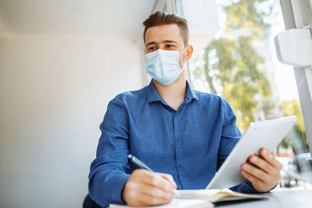 Young businessman working remotely from cafe on a laptop and tablet with a cup of coffee. Leading business during covid-19 pandemic quarantine and staying healthy conceptの写真素材