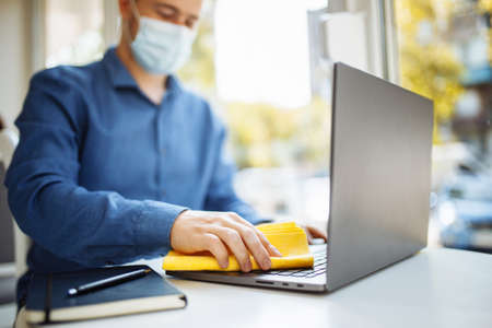 Young businessman cleans the laptop in a cafe with a yellow rag and a sanitizer to prevent coronavirus spread during worldwide pandemic. Healthcare and disinfection conceptの写真素材