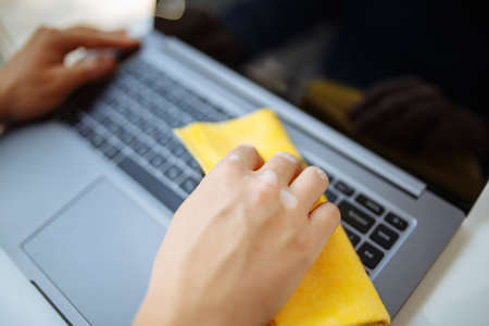 Closeup of the man's hands cleaning laptop's keyboard with a yellow rag and sanitizer to prevent...