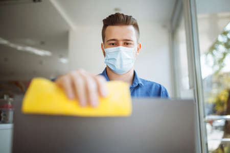 Young businessman cleans the laptop in a cafe with a yellow rag and a sanitizer to prevent coronavirus spread during worldwide pandemic. Healthcare and disinfection conceptの写真素材